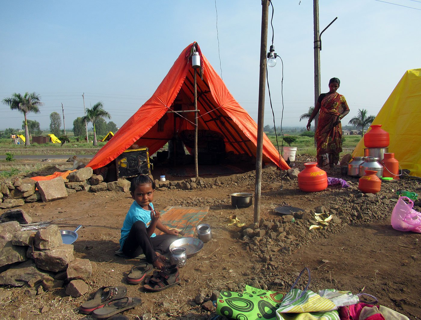 Young girl sitting outside a makeshift tent as a woman looks on in the background